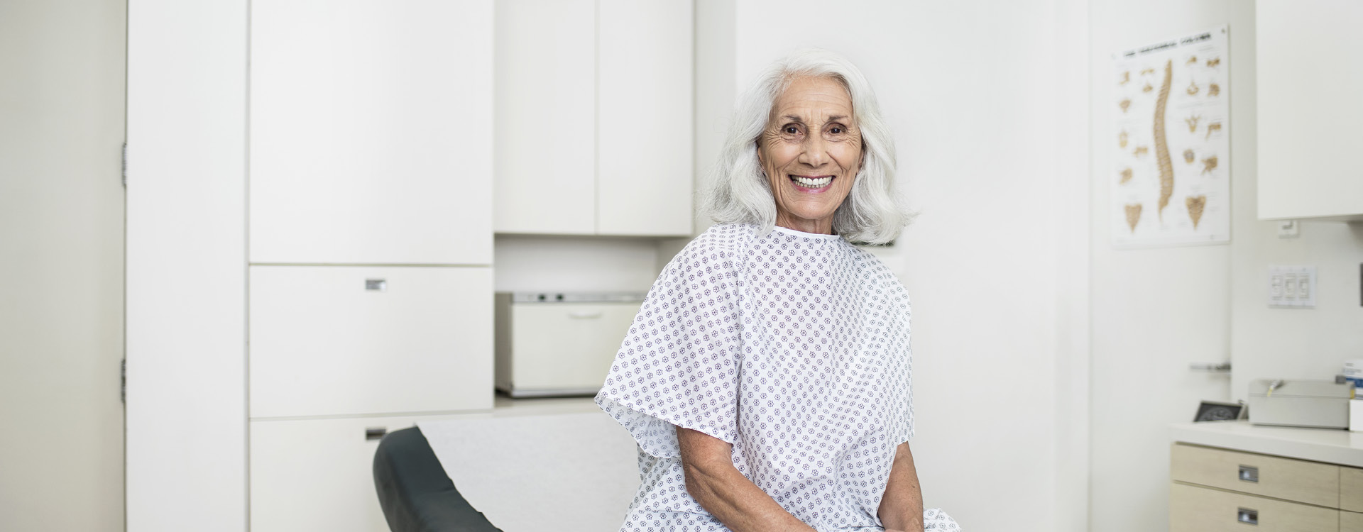 Ältere Frau mit langem hellgrauen Haar sitzt lächelnd in einem Behandlungszimmer.