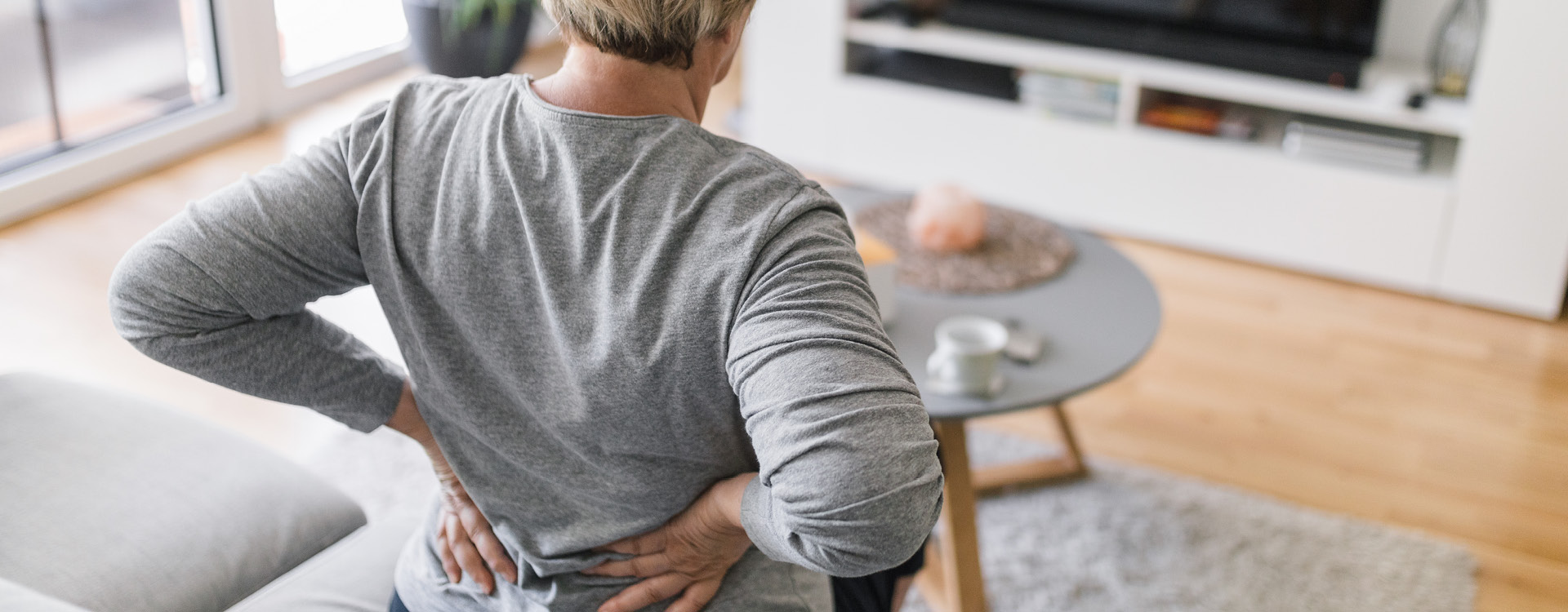 novartis-klinische-forschung-gut-informiert-gesundheitswissen-rueckenschmerzen Eine Person fasst sich mit beiden Händen an den unteren Rücken in einem Wohnzimmer.