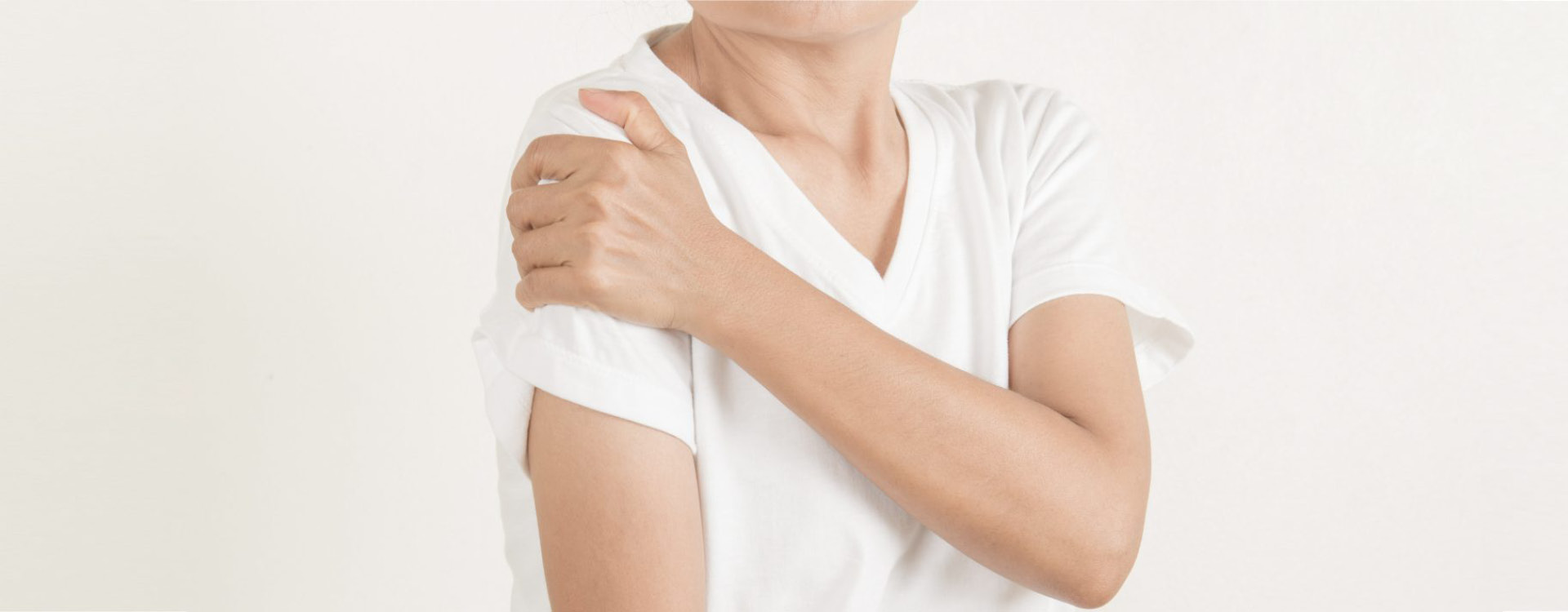 Eine Person fasst sich mit der Hand an die Schulter hellem, neutralem Hintergrund.