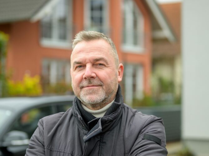 Mann in schwarzer Lederjacke mit Auto und Haus im Hintergrund | Novartis - Klinische Forschung