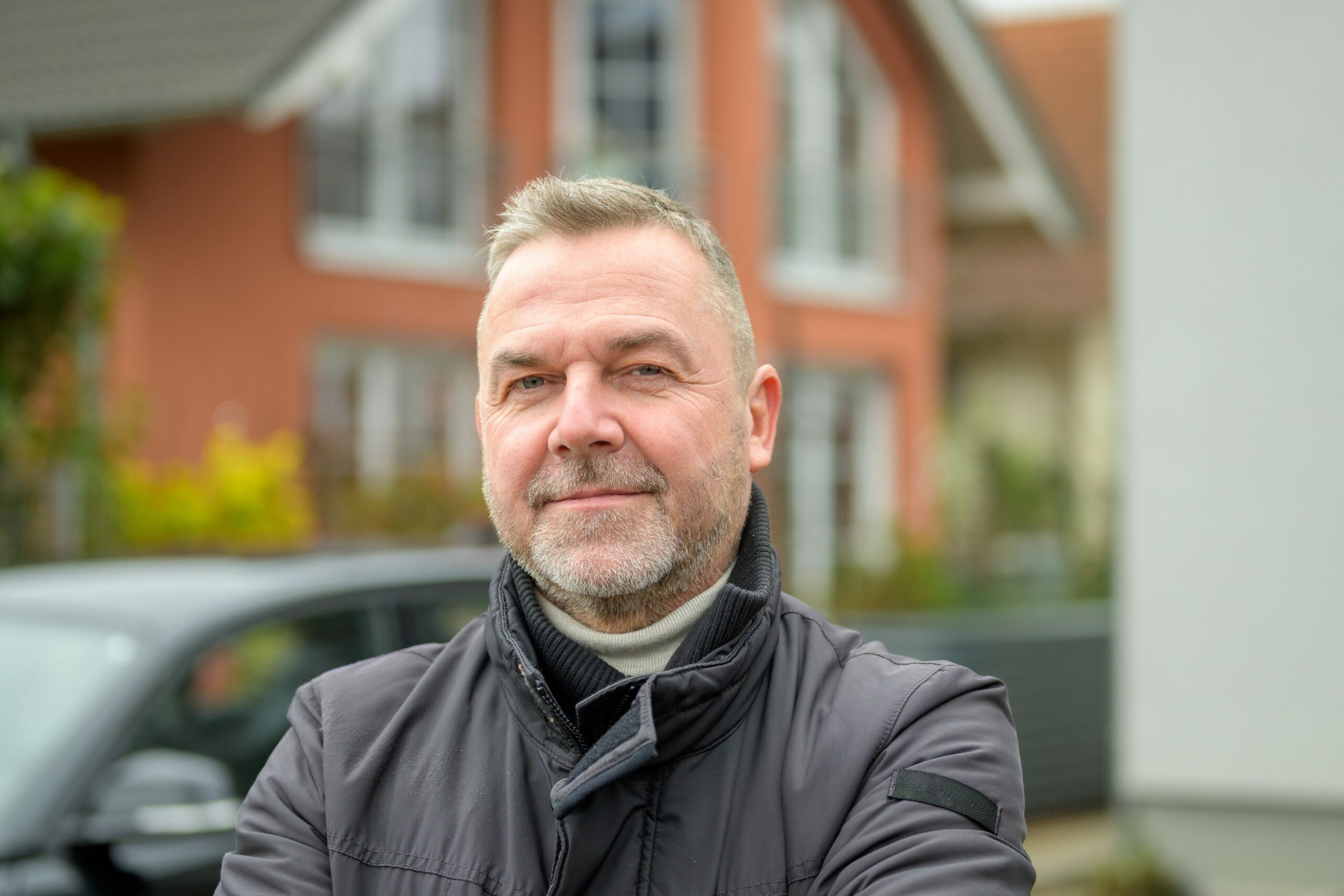 Mann in schwarzer Lederjacke mit Auto und Haus im Hintergrund | Novartis - Klinische Forschung