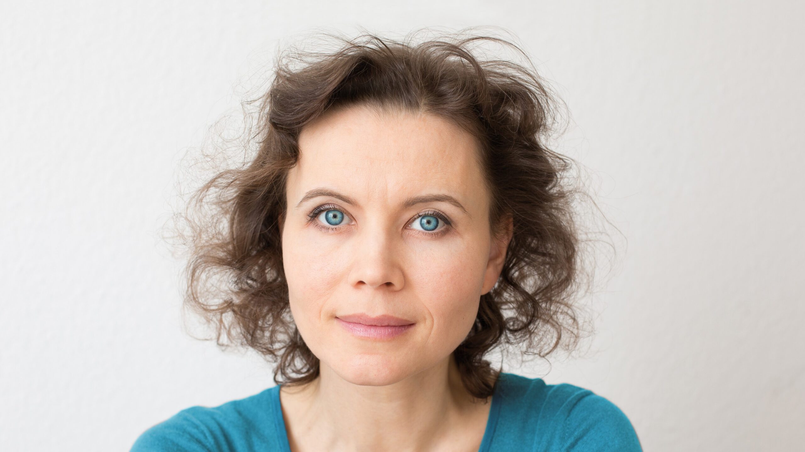 Frau mit lockigem braunen Haar und blauen Augen vor hellem, neutralem Hintergrund.