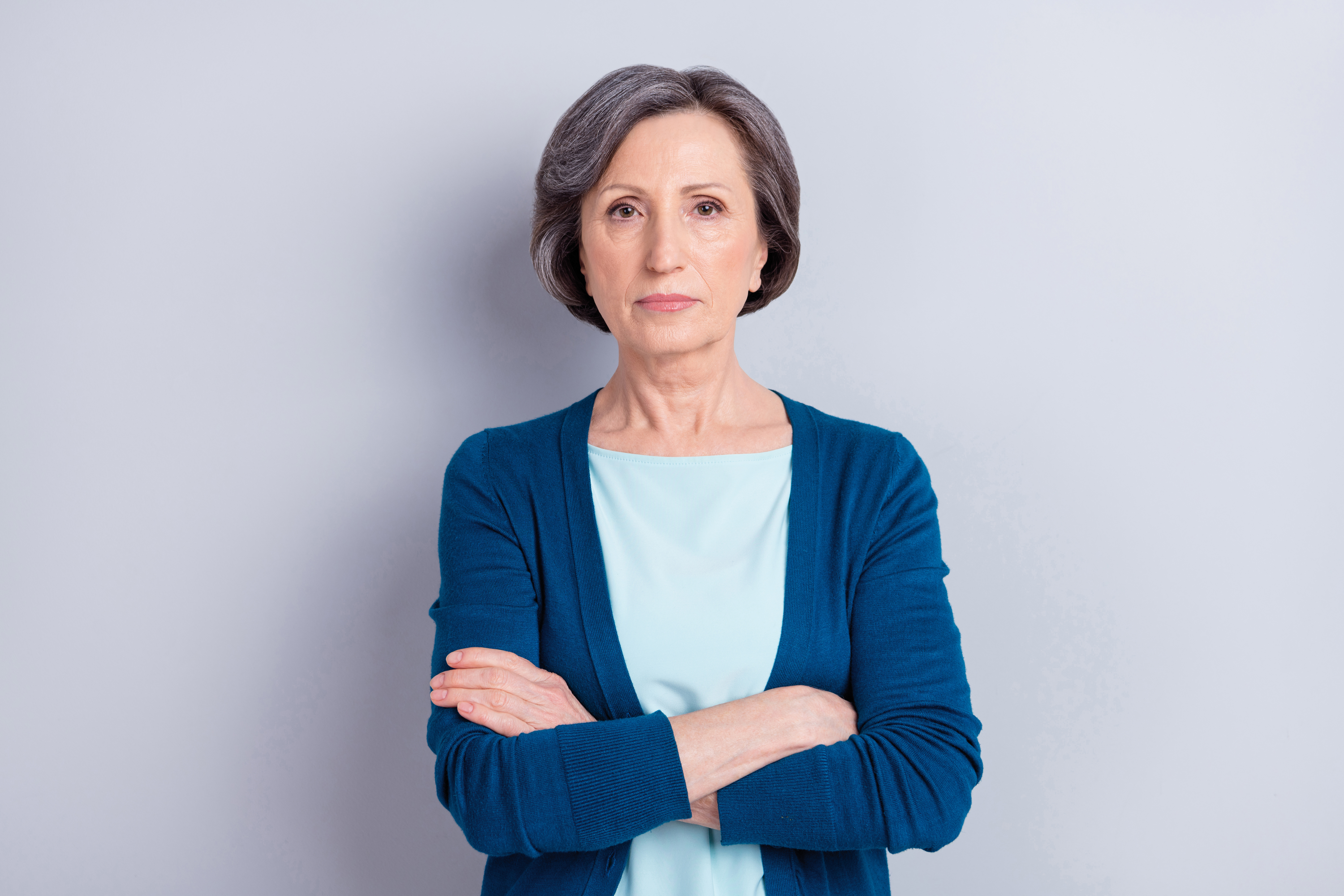 Ältere Frau mit kurzem grauem Haar verschränkt die Arme vor hellem, grauem Hintergrund.