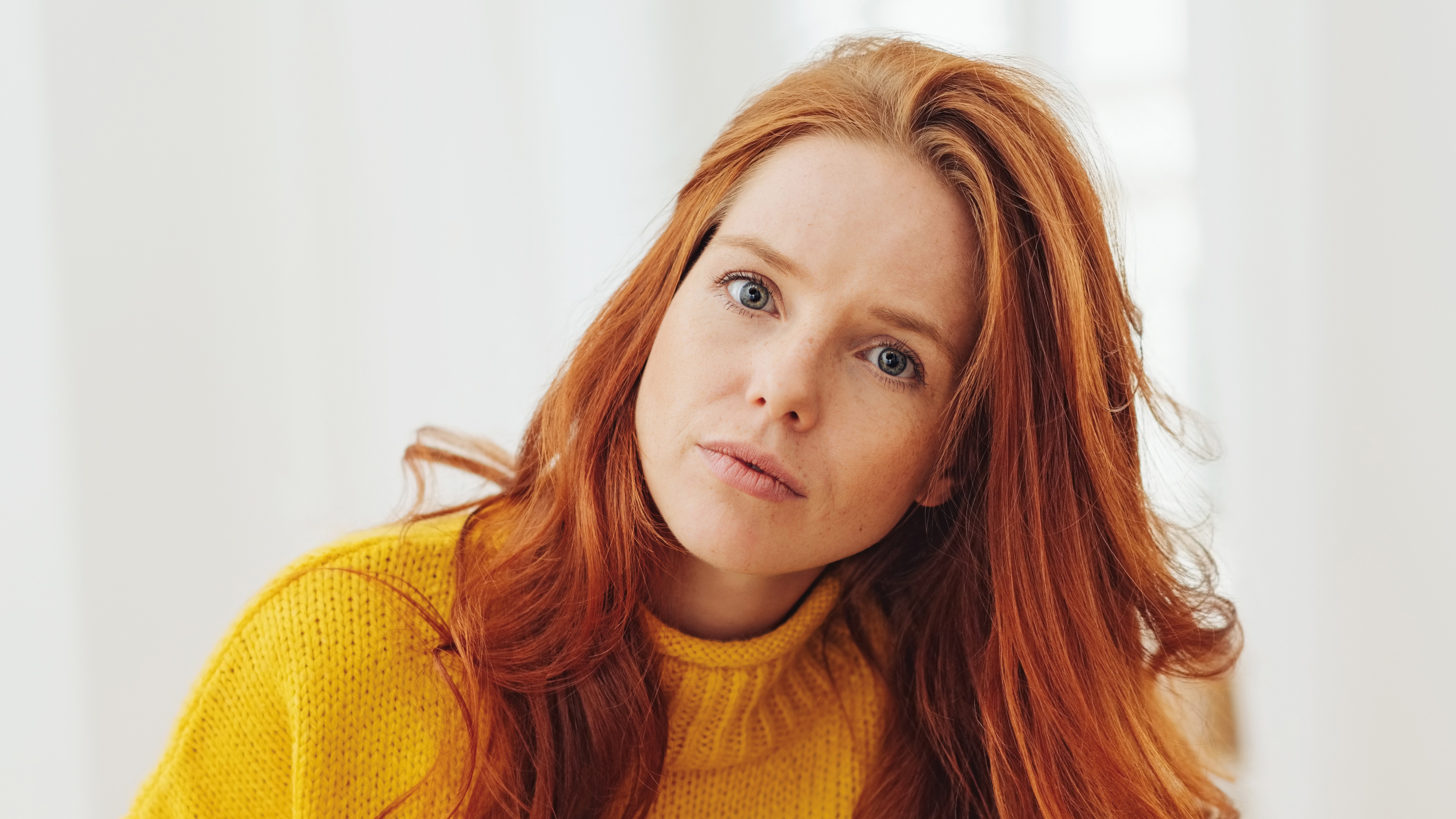 Frau mit rotem Haar und gelbem Pullover vor hellem, neutralem Hintergrund.