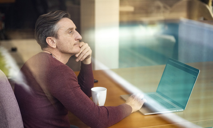 Mann sitzt am Laptop am Esstisch und blickt in die Ferne