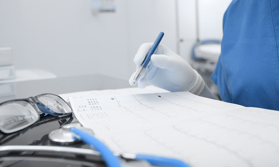 Hand einer Person in medizinischer Kleidung die ein Karidogramm an einem Schreibtisch auswertet